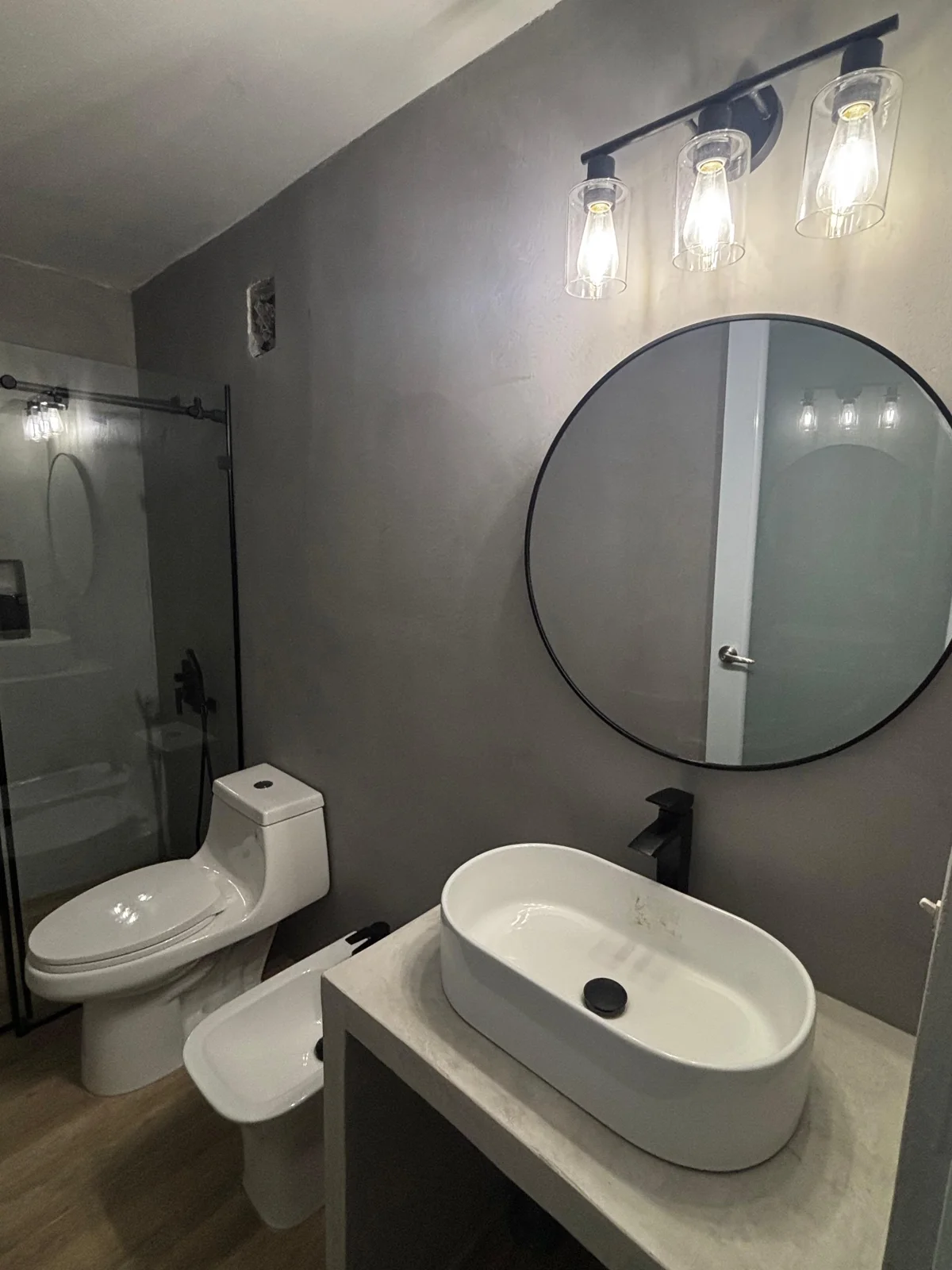 Baño con paredes grises, espejo redondo y lavamanos blanco
